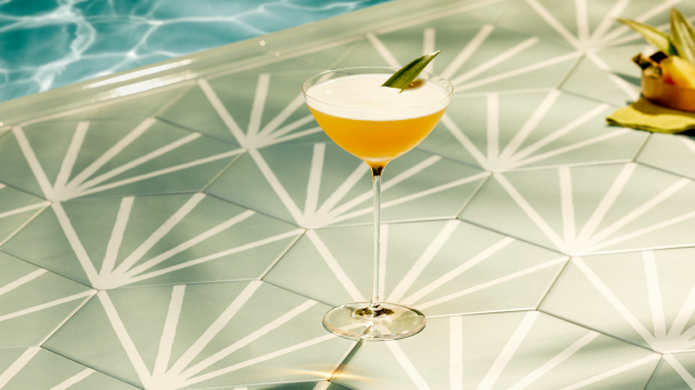 Golden cocktail in coupe glass with frothy top, green leaf garnish, placed near pool on patterned tiles.