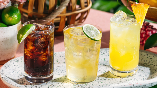 Three cocktails on tray: dark cola-like drink, light yellow cocktail, bright yellow drink with pineapple.