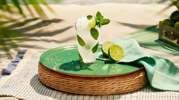 Mojito cocktail in tall glass with mint leaves, lime wedge, crushed ice, accompanied by lime halves.