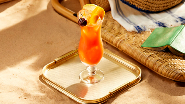 Orange cocktail in hurricane glass with orange slice, cherry, placed on golden tray near beach accessories.