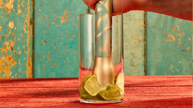 Hand using gold and teal muddler to press lime wedges in clear glass.