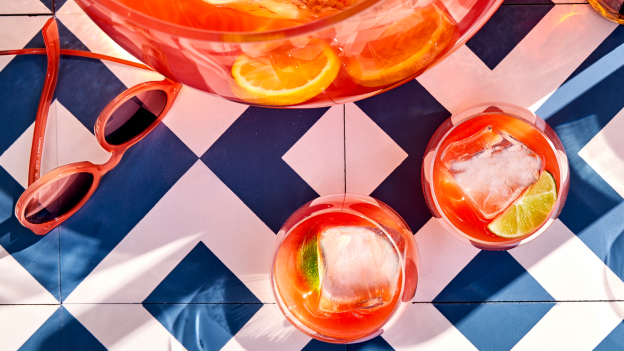 Overhead view of punch bowl, two glasses with red cocktail, citrus slices, sunglasses on checkered surface.