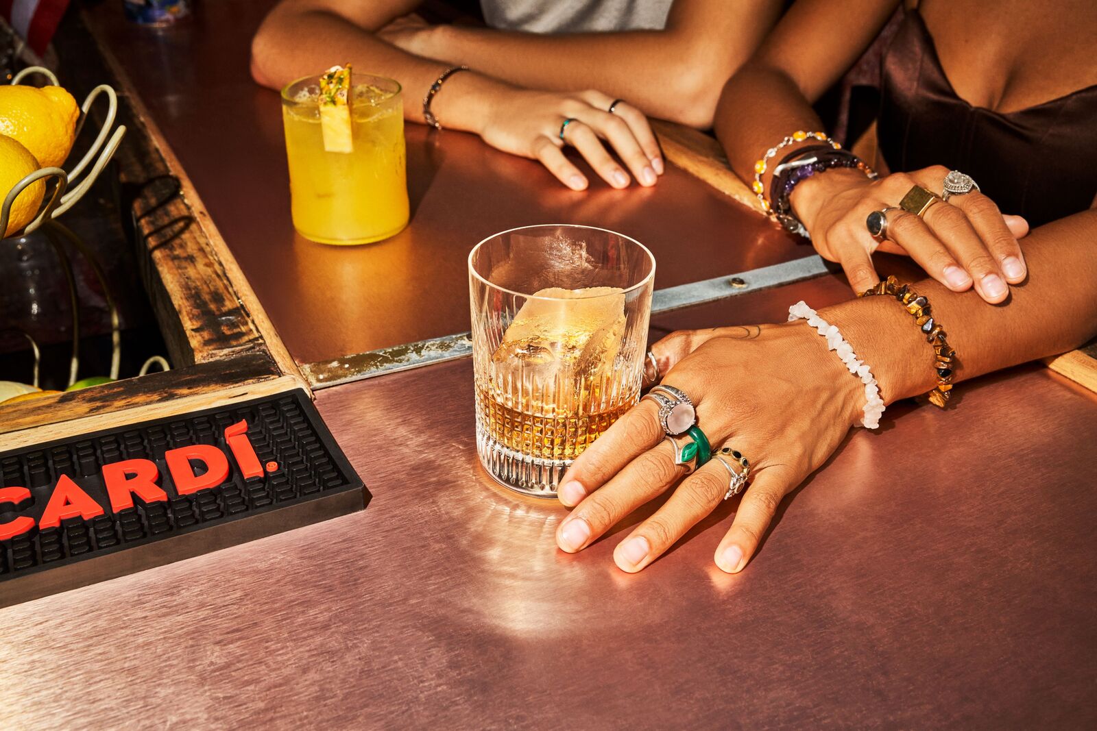 Two cocktails on a bar: amber drink with large ice cube, yellow cocktail with pineapple garnish.