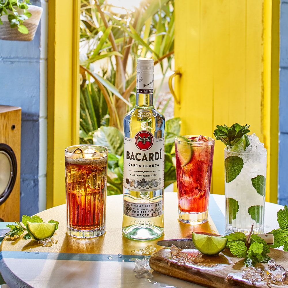 Bacardí Carta Blanca rum bottle with three cocktail glasses, lime wedges, mint leaves scattered around.