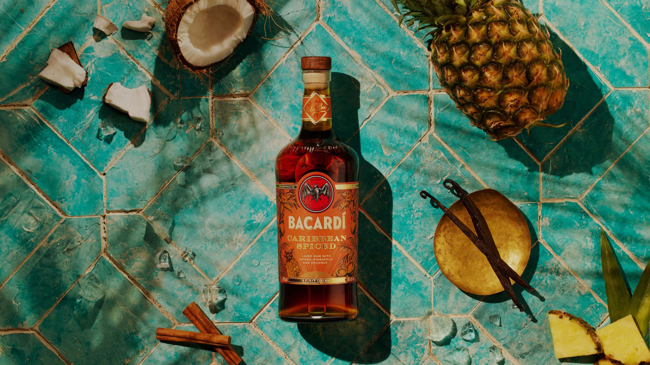 BACARDÍ Caribbean Spiced rum bottle surrounded by pineapple, coconut, cinnamon sticks, vanilla beans, ice cubes.