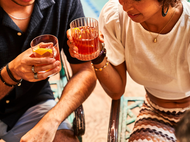 Two people holding cocktails with orange garnish in an outdoor setting.