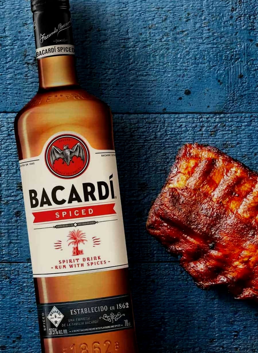 BACARDÍ Spiced rum bottle next to a piece of grilled or roasted meat with caramelized crust.