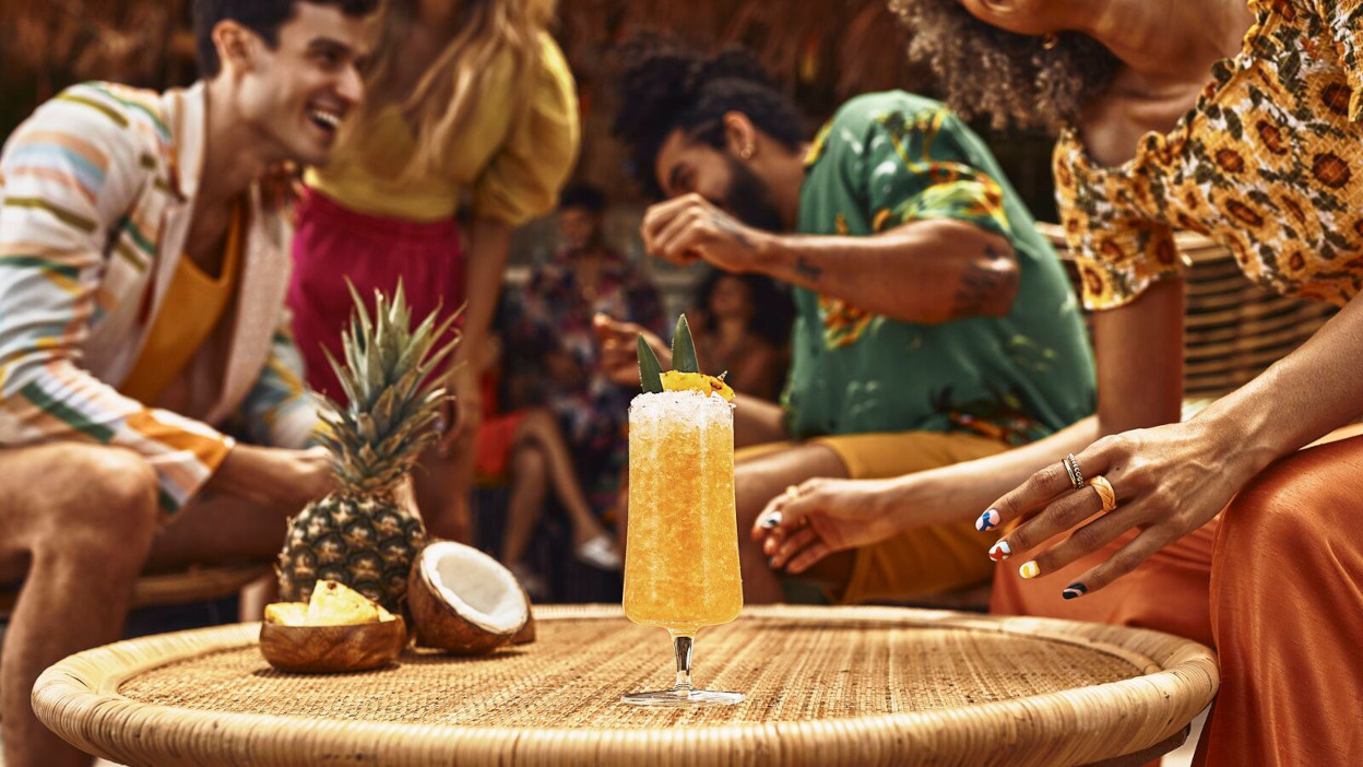 Tropical cocktail with pineapple garnish on rattan table, surrounded by pineapple, coconut, people.