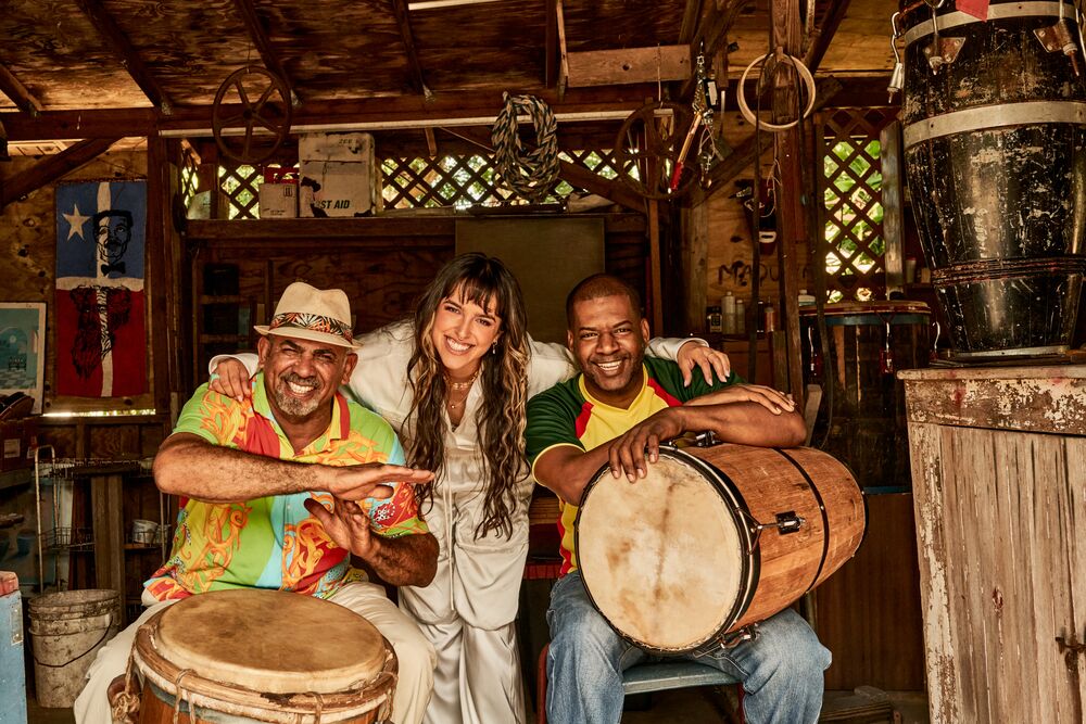 Three musicians posing together with drums in a rustic setting, smiling and closely positioned.