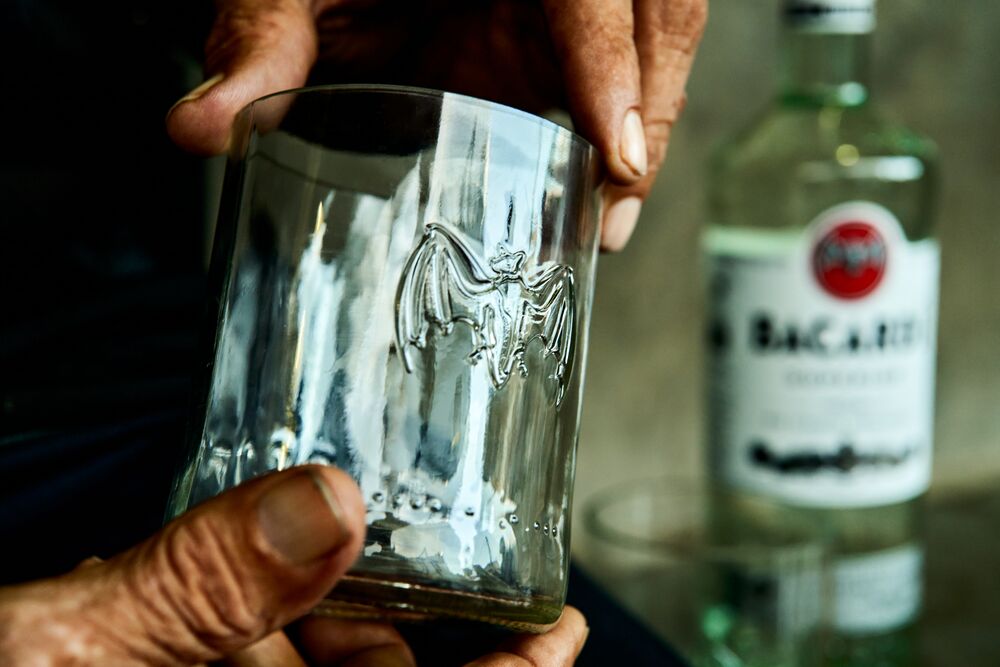 Hands holding a clear glass tumbler with embossed BACARDÍ bat logo, BACARDÍ bottle visible in background.