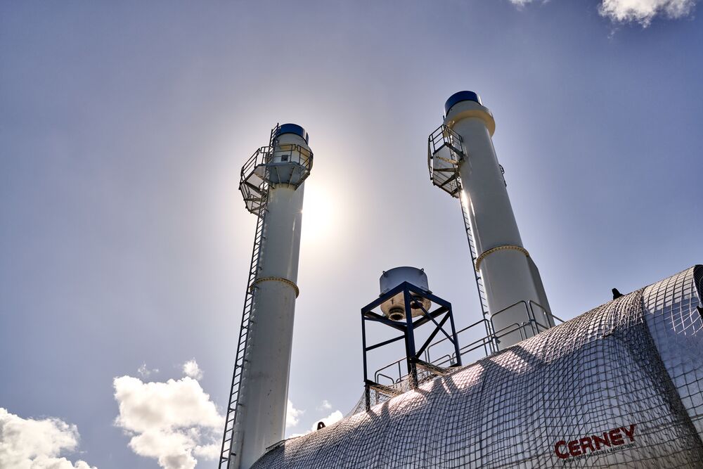 Two tall white industrial chimneys with blue caps, silhouetted against sunny sky with curved industrial structure.