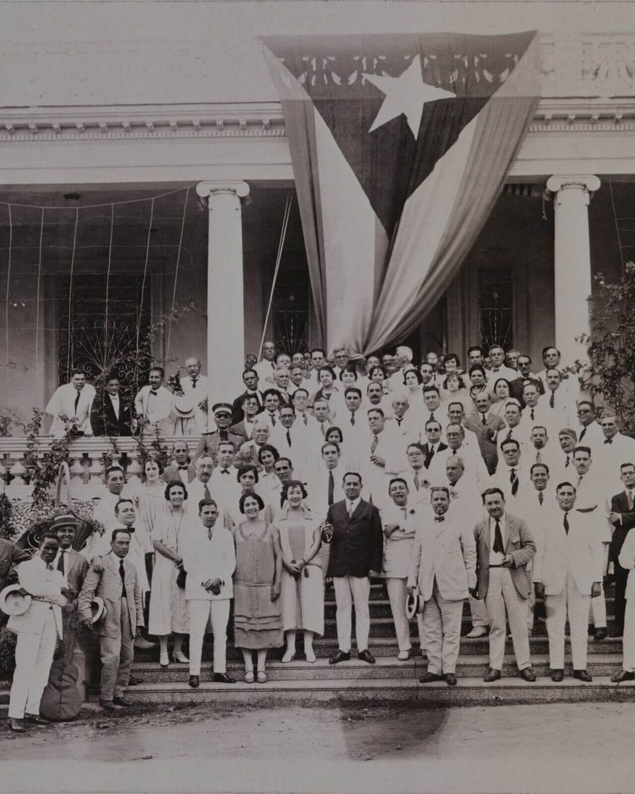 Large group of people posing on building steps with Cuban flag draped from balcony.