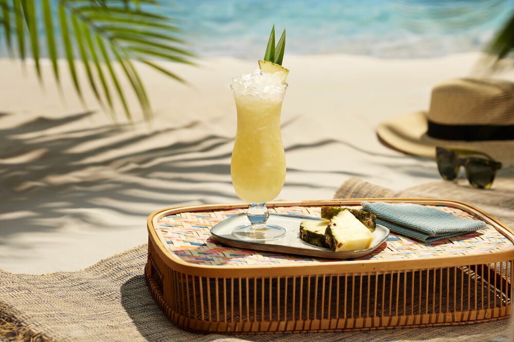 Tall yellow cocktail with crushed ice and pineapple garnish on a woven tray.