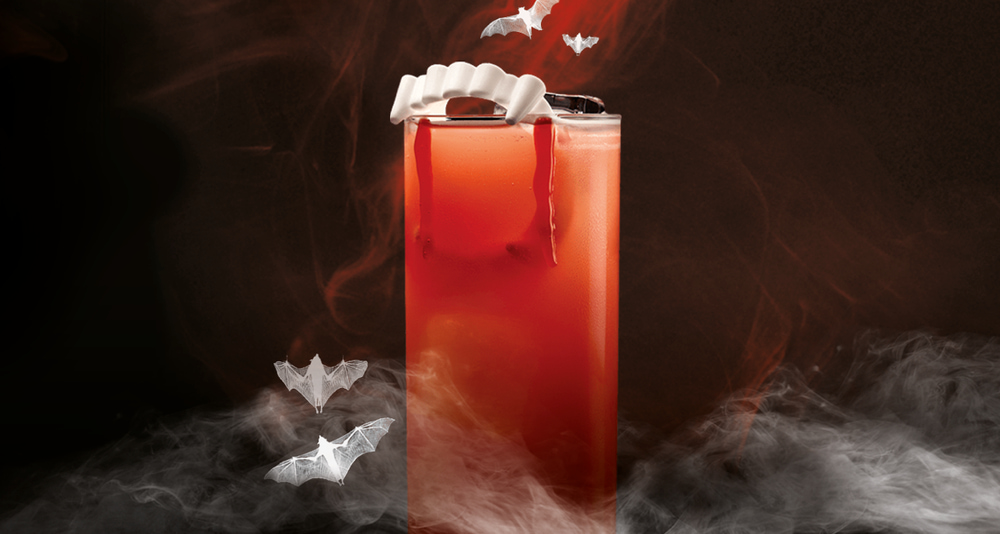 Red cocktail with vampire teeth garnish and blood-like syrup drips, surrounded by paper bats.