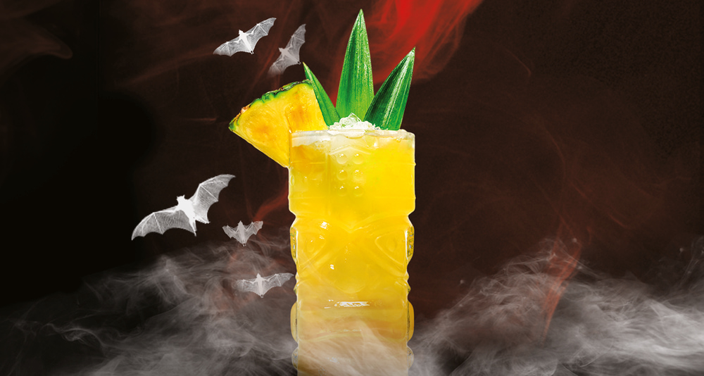 Yellow tiki-style cocktail garnished with pineapple, mint leaves, surrounded by white bats.