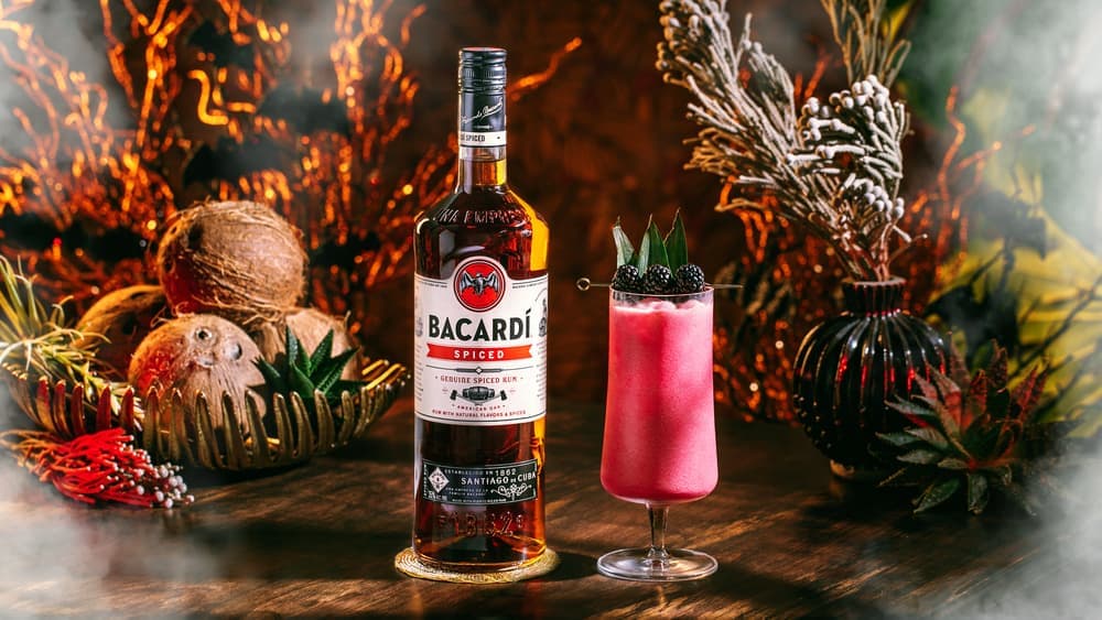 BACARDÍ Spiced rum bottle with pink cocktail, garnished with blackberries, pineapple leaves.