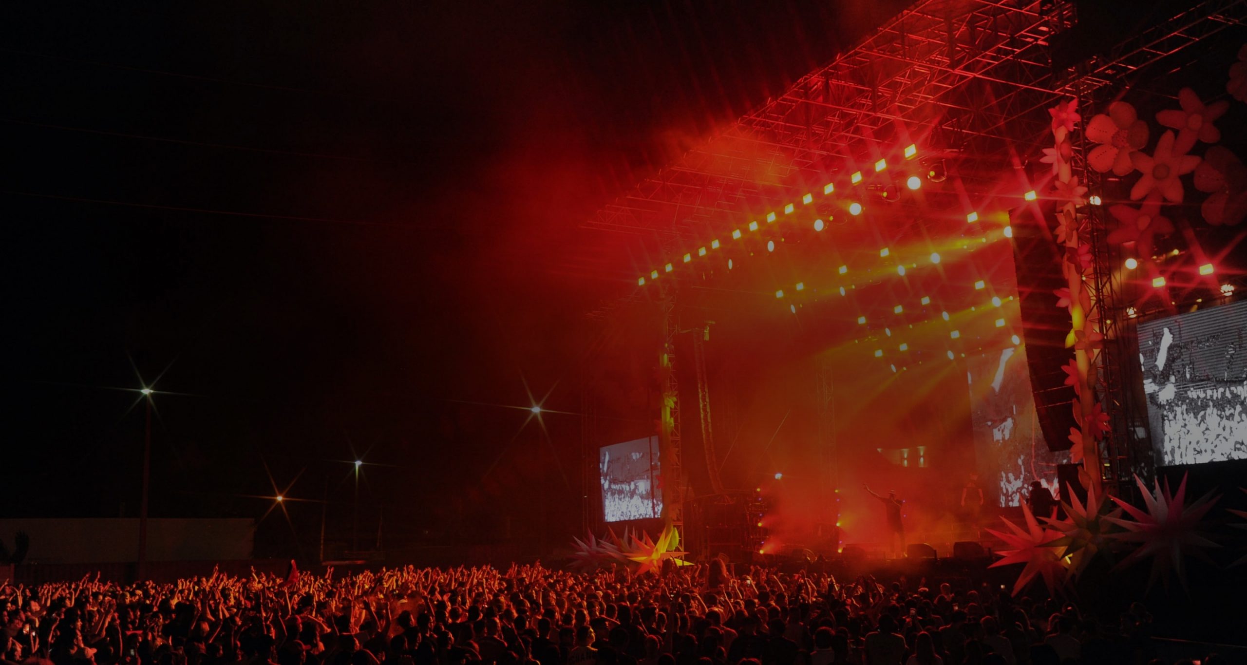 Nighttime outdoor concert with massive crowd, illuminated stage, vibrant red and orange spotlights.