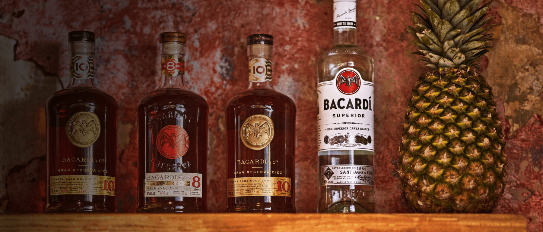 Four BACARDÍ rum bottles with whole pineapple arranged on wooden shelf.