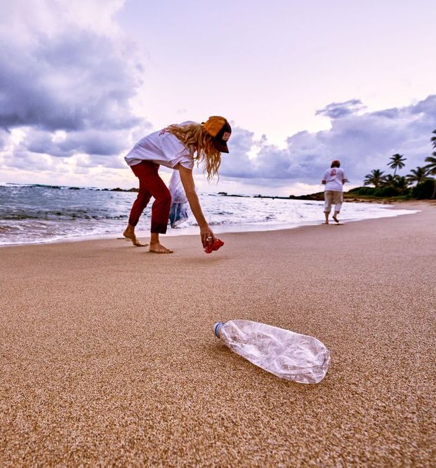 Person bending to collect trash from sandy beach with plastic bag and bottle.