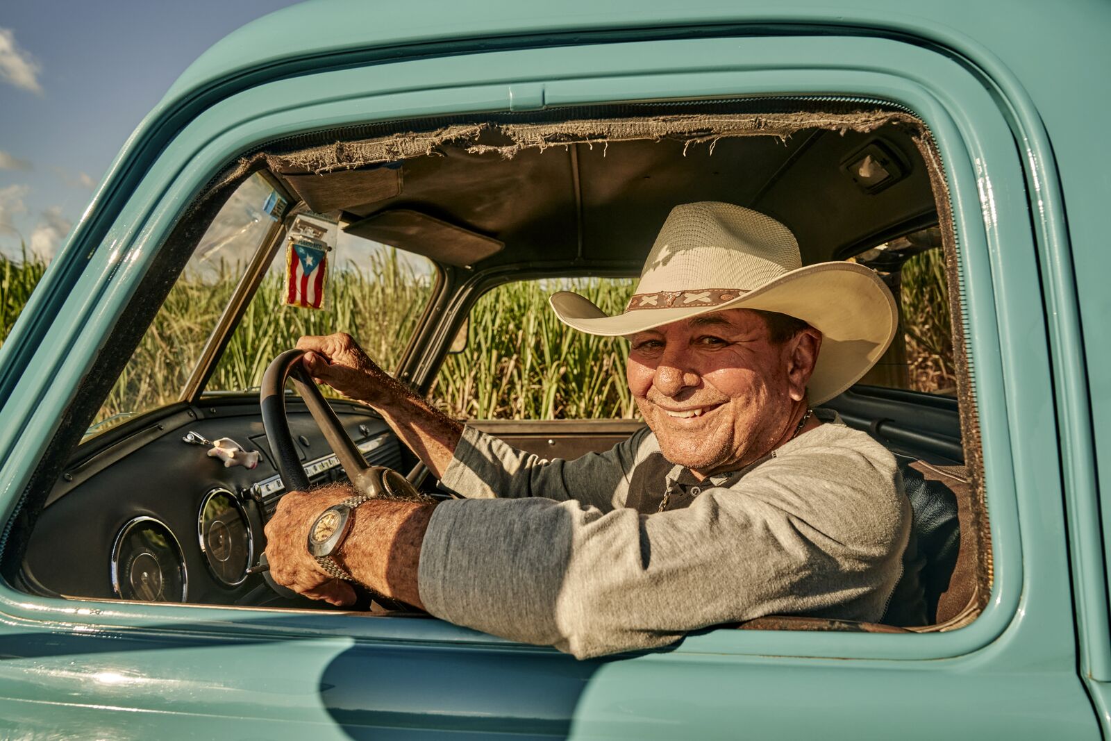 Person wearing a cowboy hat smiling while driving an old turquoise truck with sugarcane field visible through windows.