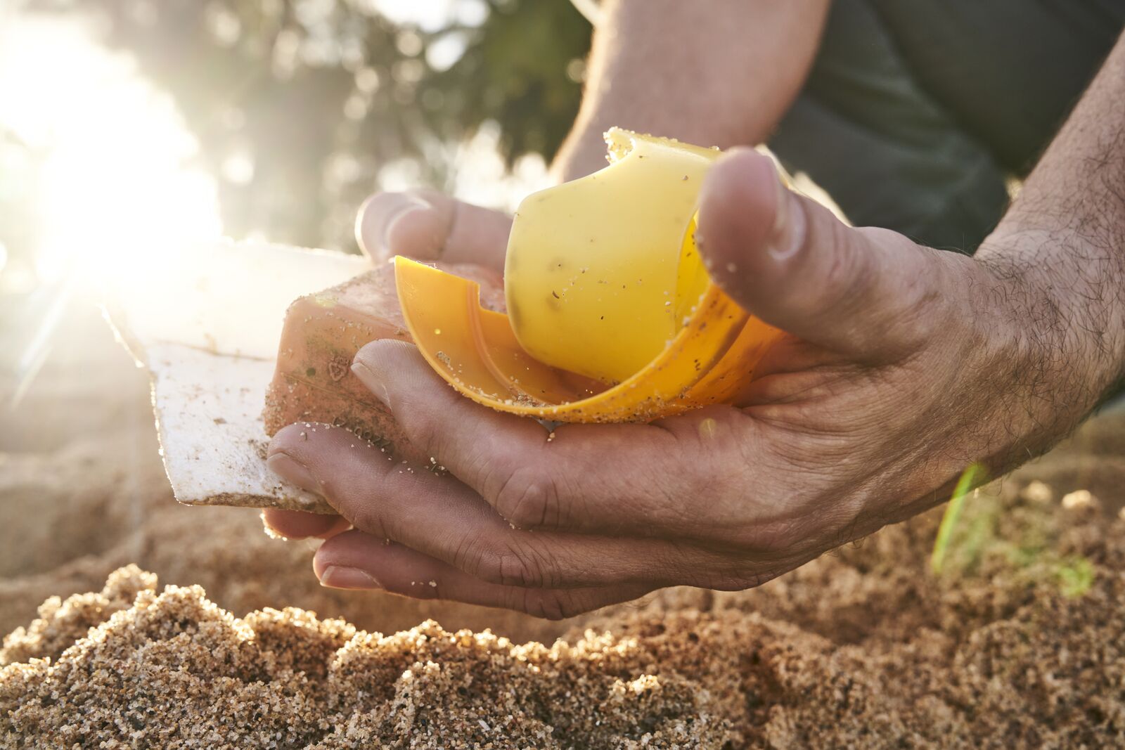 Hands cupped over sandy ground, holding pieces of yellow and white plastic debris.