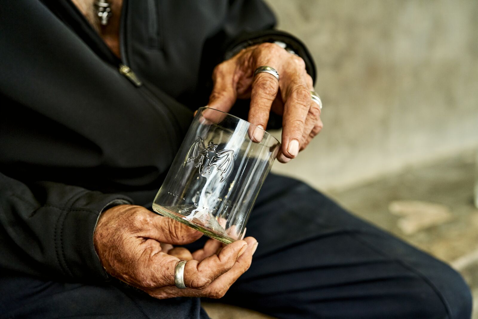 Hands holding a glass with etched BACARDÍ bat logo, adorned with silver rings.