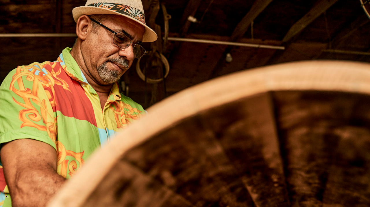 Person in a colorful shirt and fedora hat working intently with a large wooden barrel.
