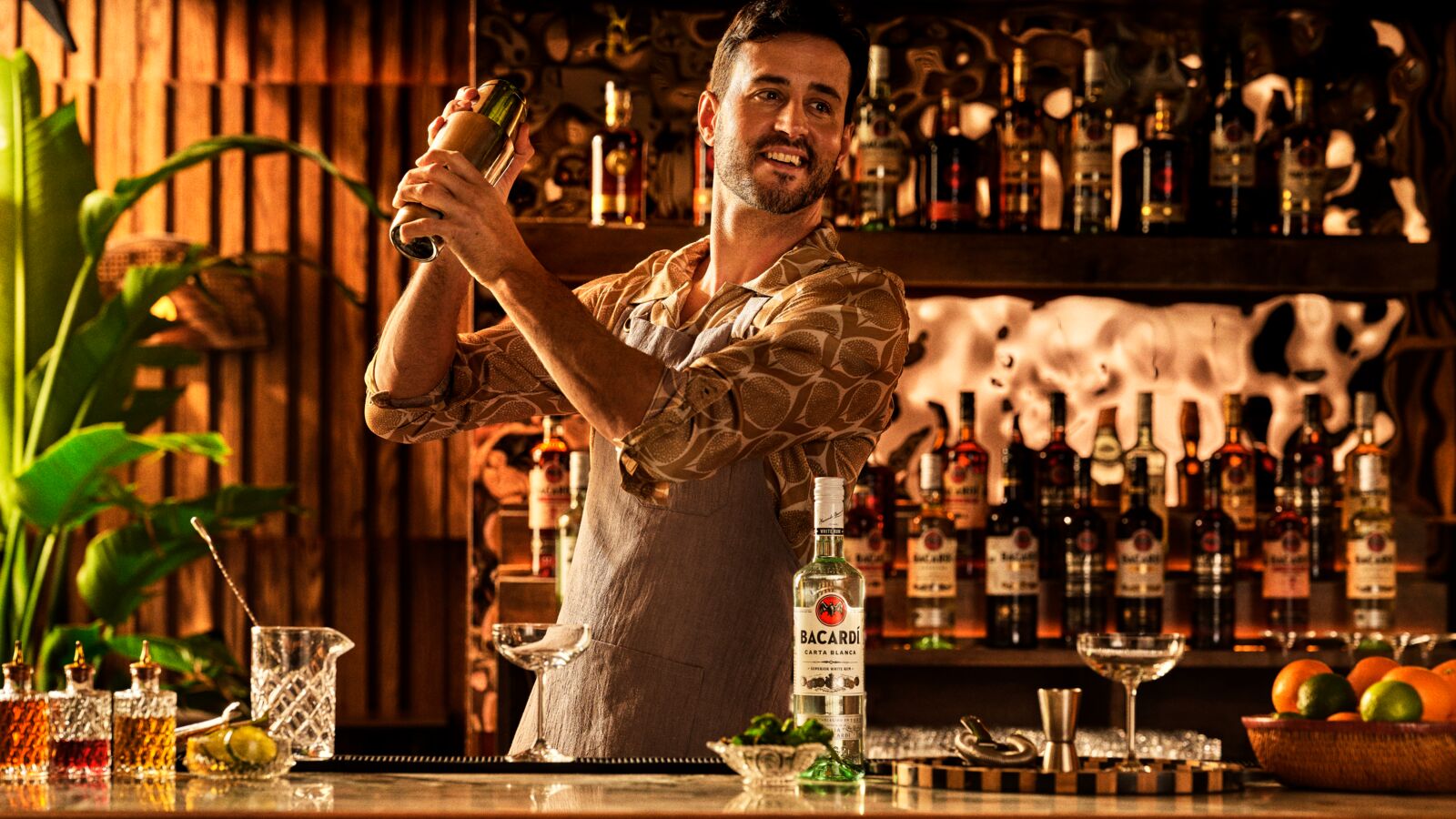 Bartender in an apron shaking a cocktail behind a bar with BACARDÍ bottles.