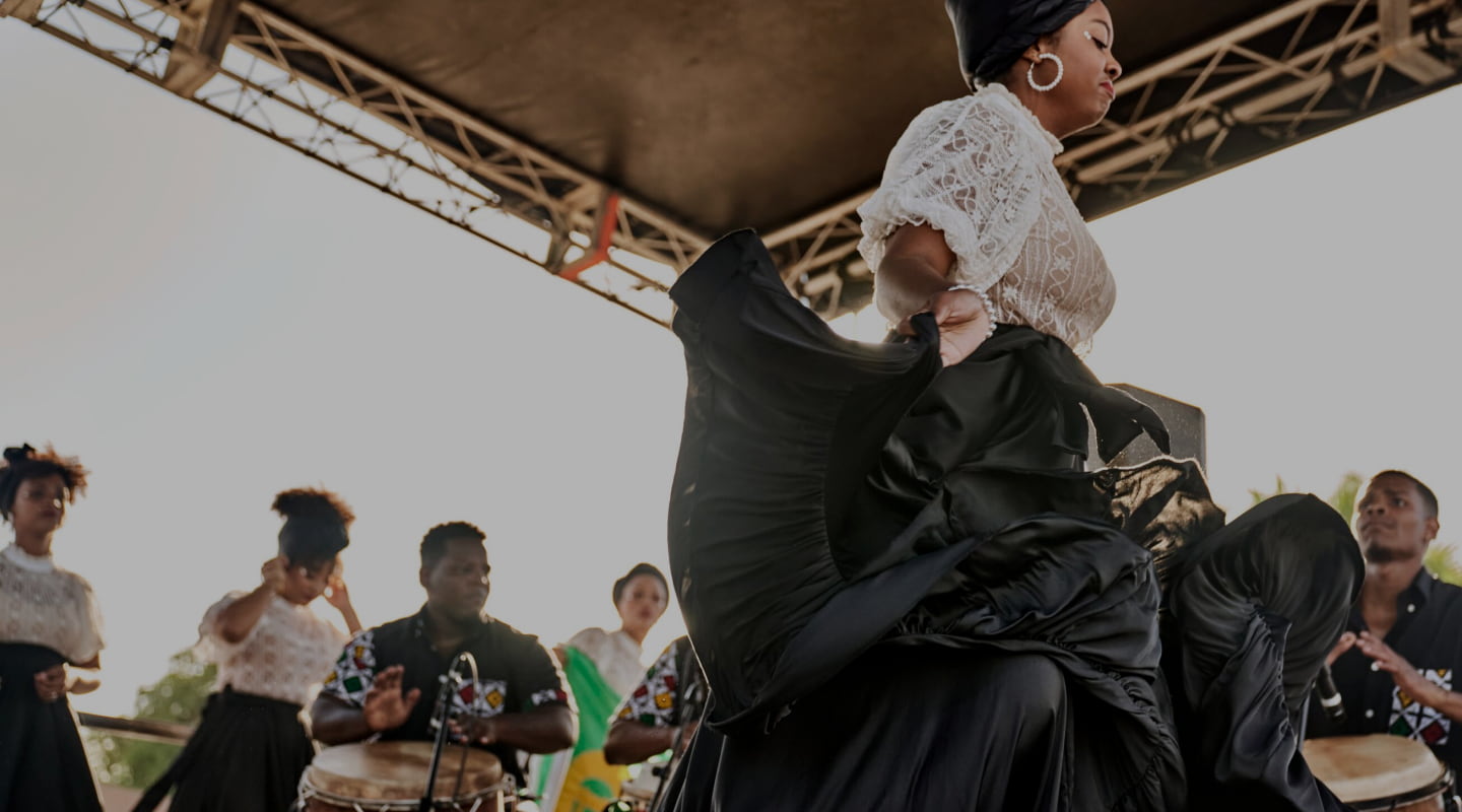 Dynamic dancer performing on an outdoor stage with drummers in the background.