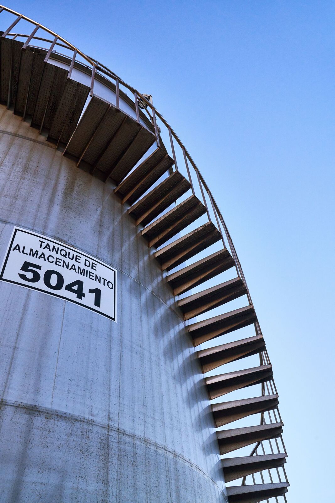 Grey industrial storage tank with metal spiral staircase winding along its curved surface.