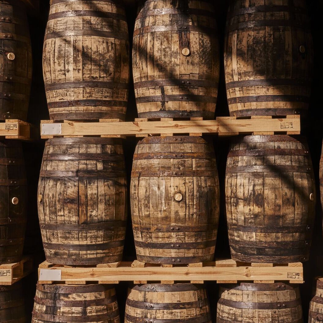 Stacked wooden barrels with rusty metal bands on wooden pallets in dimly lit environment.