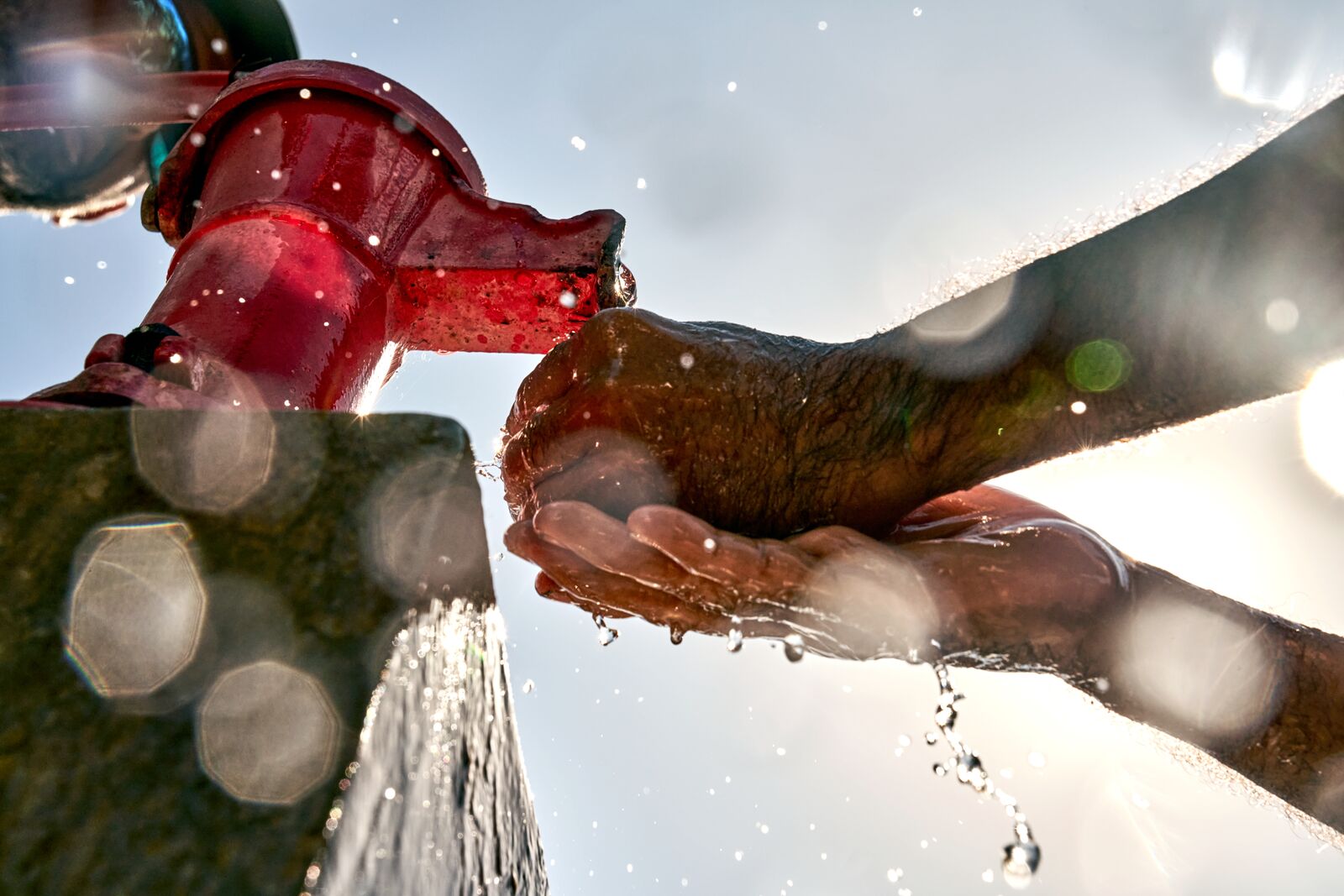 Hands cupped beneath a red faucet, catching clear water droplets sparkling in bright sunlight.