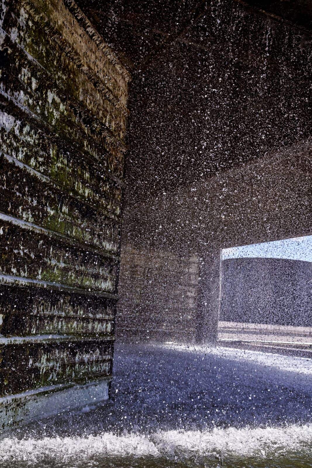 Water splashes and sprays within a textured concrete structure with moss-covered walls.