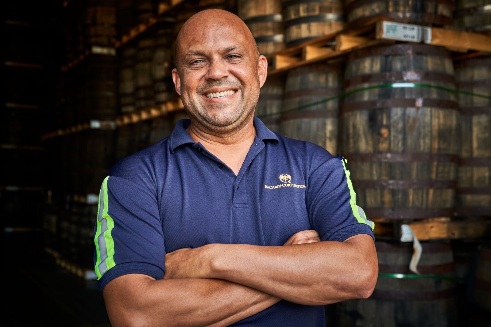 Employee standing with crossed arms in a distillery with wooden barrels in the background.
