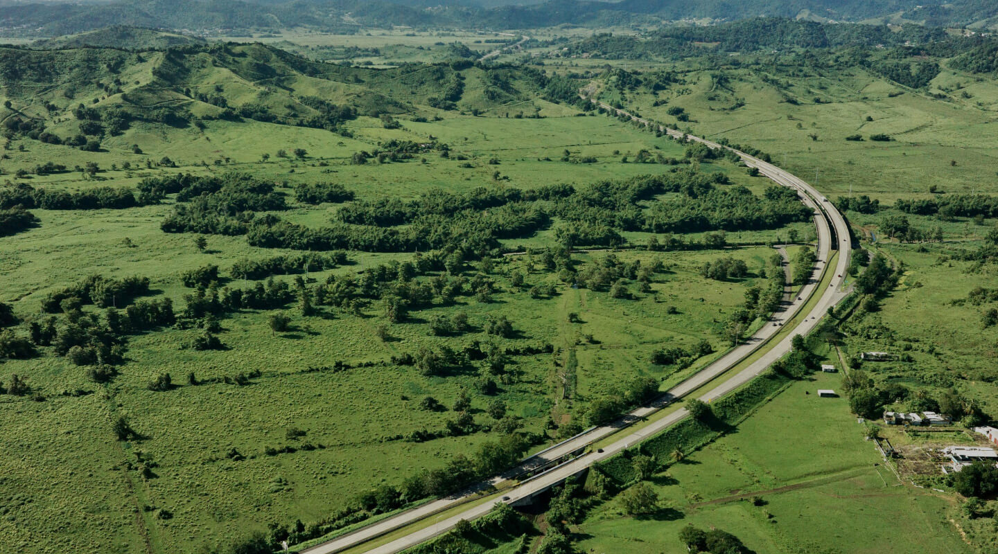 Aerial view of a highway curving through verdant rolling hills with scattered trees and small buildings.