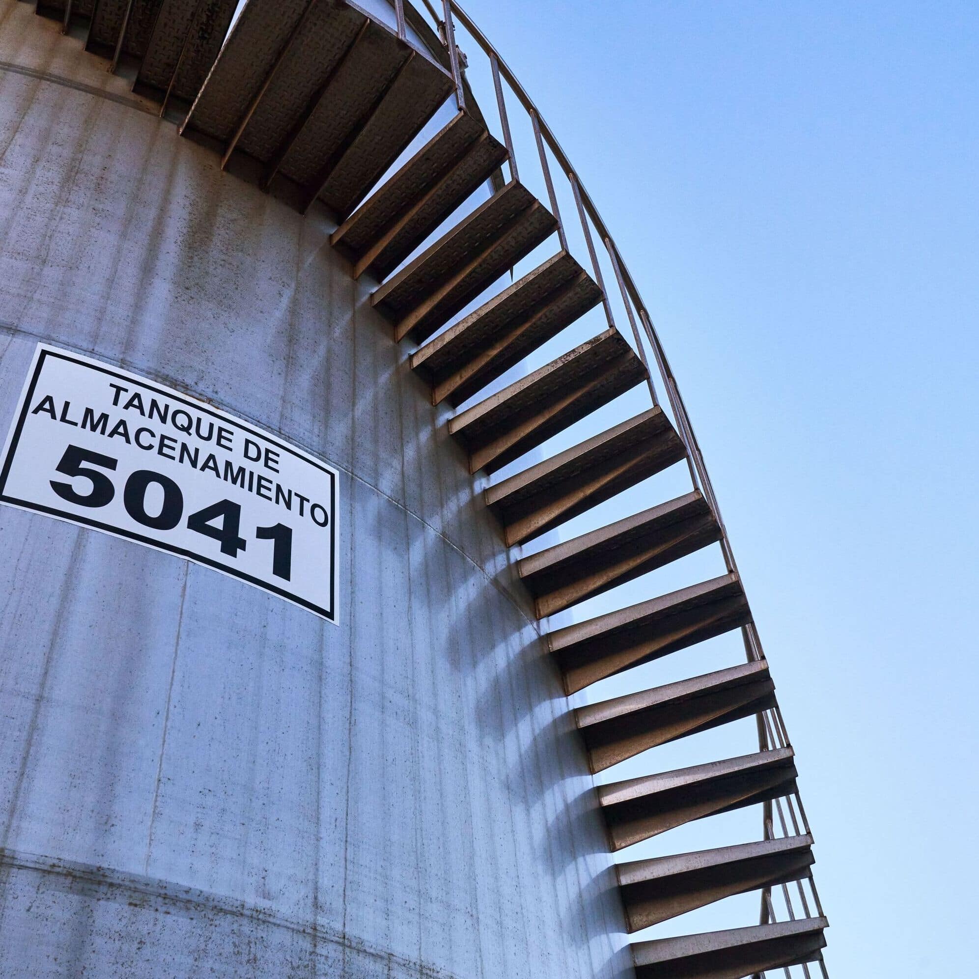 Cylindrical industrial storage tank with spiral metal staircase, marked with 'TANQUE DE ALMACENAMIENTO 5041' sign.