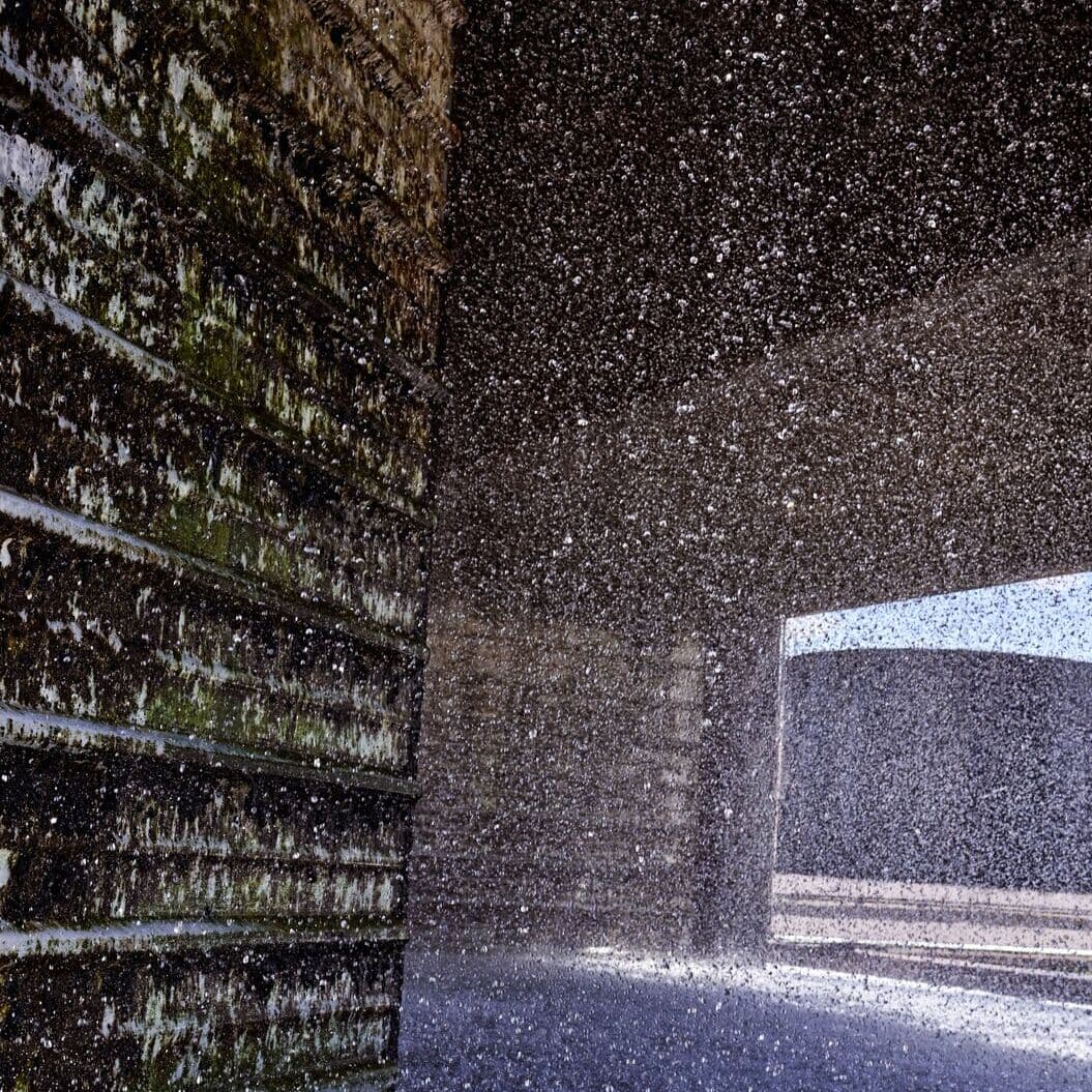 Water droplets suspended in air, illuminated by bright light near textured wall with moss.