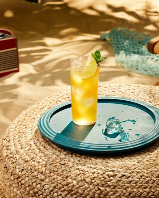 Yellow cocktail in tall glass with lime wedge and mint, placed on a teal tray.