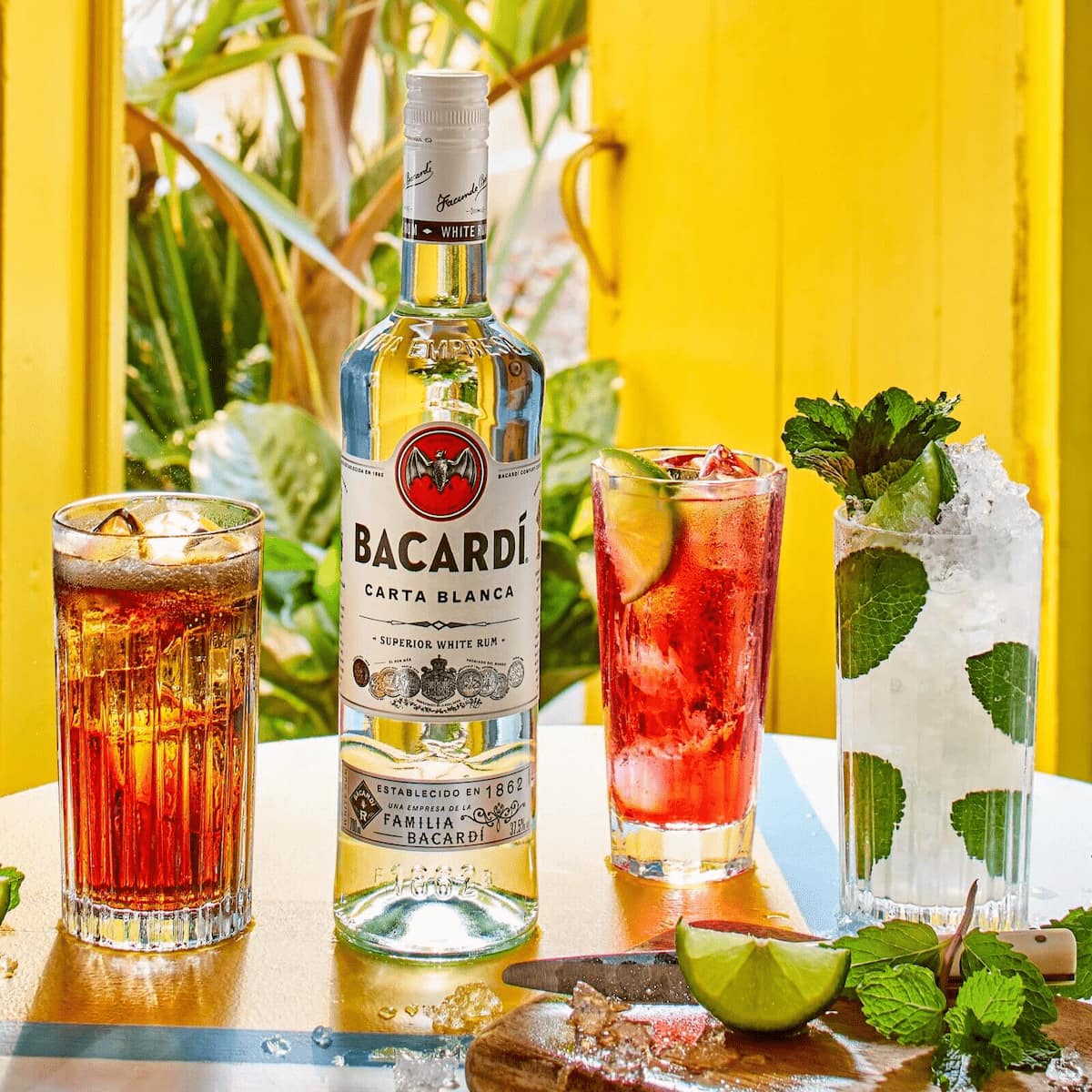 BACARDÍ Carta Blanca rum bottle with three cocktails, lime wedges, mint leaves.