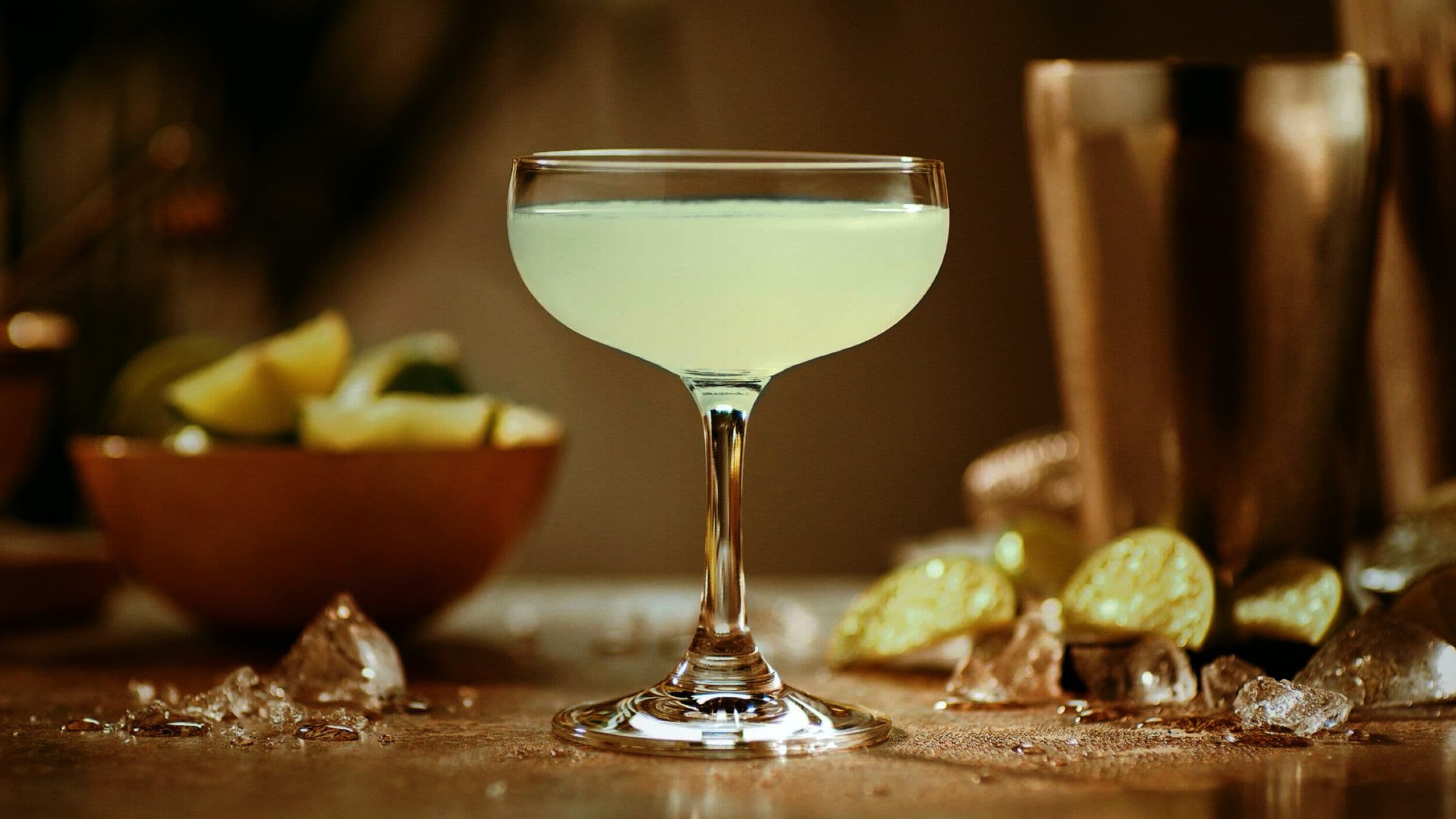Pale green Daiquiri in a coupe glass with lime wedges, cocktail shaker, scattered ice cubes.