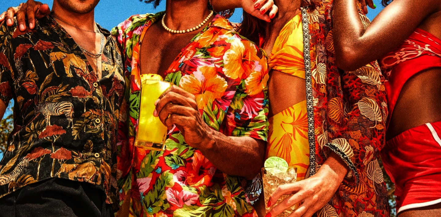 Group of people in tropical clothing holding cocktails, standing close together under bright sunlight.