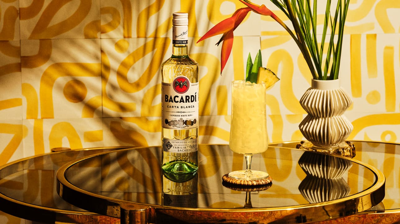BACARDÍ Carta Blanca rum bottle next to a tropical cocktail garnished with pineapple and green leaves.