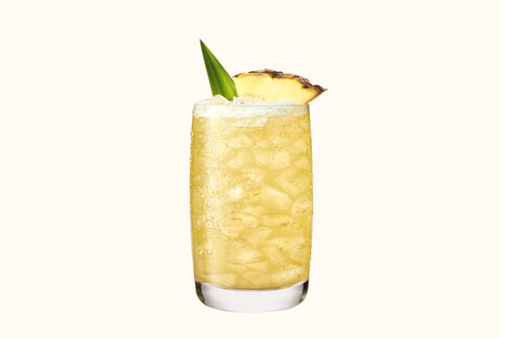 Tall glass with pale yellow cocktail, crushed ice, pineapple wedge, and green leaf with condensation.