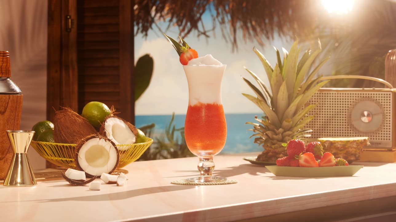 Layered tropical cocktail in hurricane glass surrounded by fresh ingredients, coconuts, limes, pineapple, strawberries.