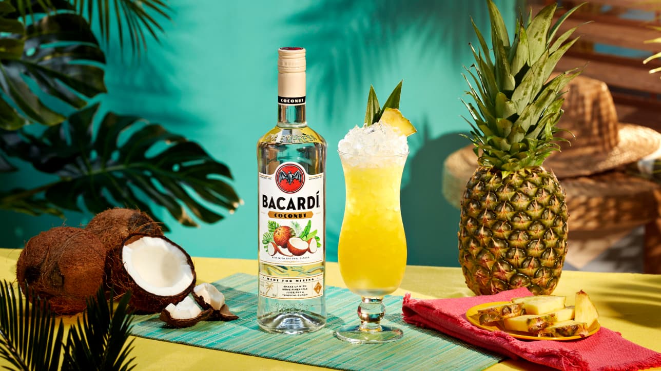 BACARDÍ Coconut rum bottle with yellow tropical cocktail, surrounded by coconuts, pineapple slices.