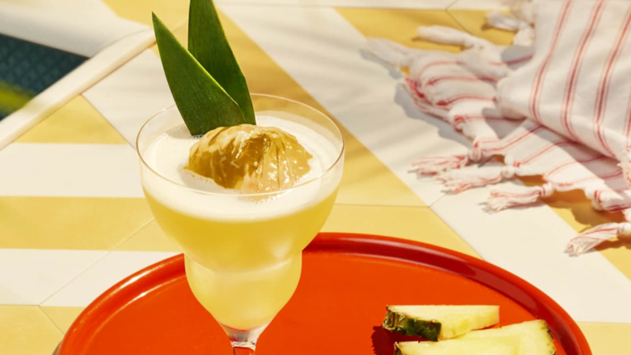 Yellow cocktail in stemmed glass with pineapple leaves, large ice cube, placed on an orange tray with pineapple wedges.