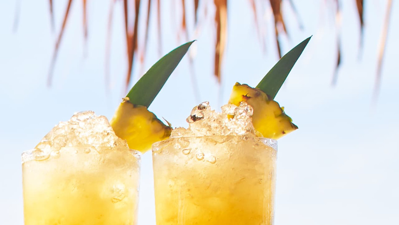 Two yellow tropical cocktails with crushed ice, pineapple wedges, green leaves near a blurred thatched roof.