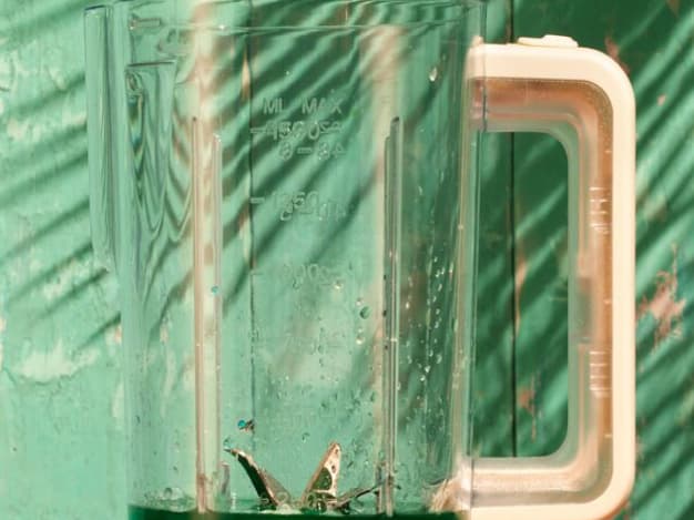 Clear plastic blender jar with dark green liquid, positioned against a weathered teal wooden wall.