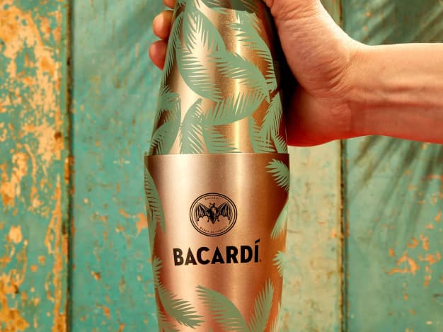 Hand holding a metallic copper cocktail shaker with green palm leaf patterns and BACARDÍ logo.