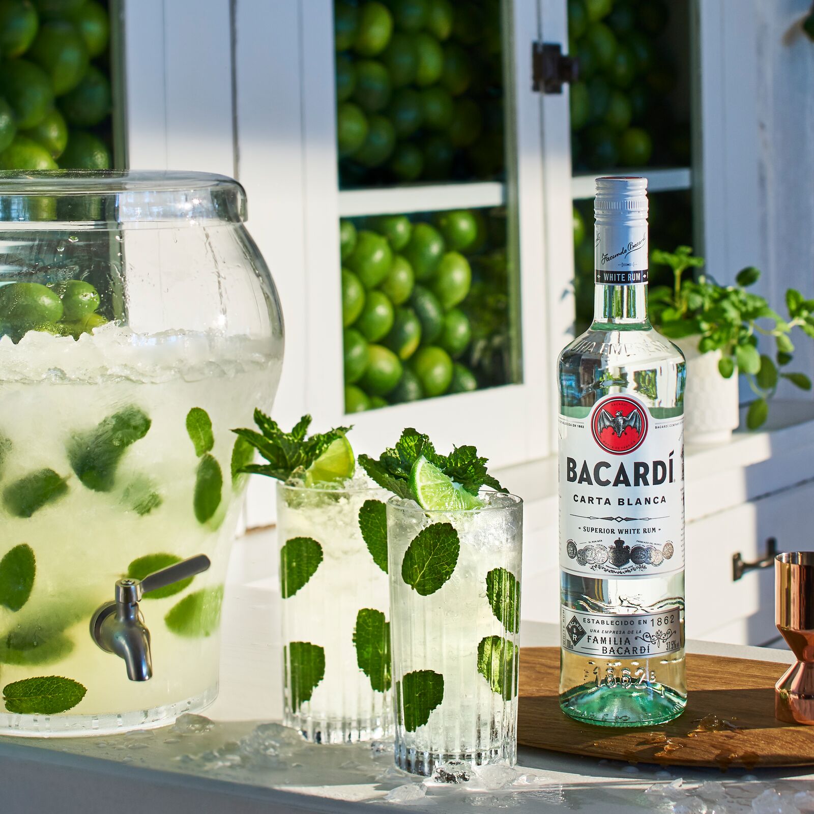 BACARDÍ Carta Blanca rum bottle beside mojito glasses, large dispenser with lime slices, mint.